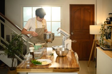 Genç Asyalı adam modern bir mutfakta çırpılmış krema yapmak için karıştırıcı kullanıyor..