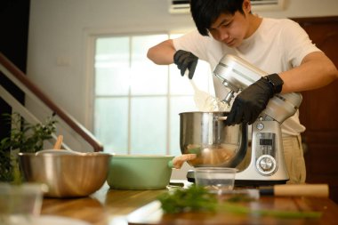 Genç Asyalı adam modern bir mutfakta çırpılmış krema yapmak için karıştırıcı kullanıyor..