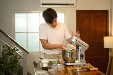 Genç Asyalı adam modern bir mutfakta çırpılmış krema yapmak için karıştırıcı kullanıyor..