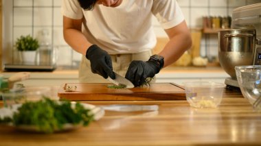 Siyah eldivenli bir adam tahta kesme tahtasına otları kesiyor..