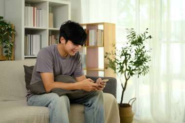 Evdeki koltukta cep telefonu kullanarak günlük giysiler içinde gülümseyen Asyalı adam..