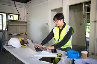 Planları gözden geçirmek ve konut yenileme çalışmalarını yönetmek için dizüstü bilgisayar kullanan mühendis.