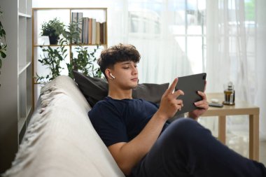 Genç adam kulaklıklarla kanepede uzanıyor ve dijital tablette içeriği izliyor..
