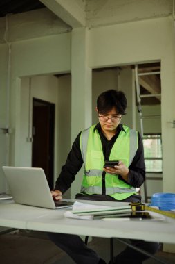 Mühendis, geçici çalışma masasındaki akıllı telefonu dizüstü bilgisayar ve planlarla kontrol ediyor..