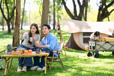 Çocuk arka planda oynarken Asyalı çift kamp alanında sıcak içeceklerin tadını çıkarıyor..