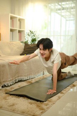 Sıcak bir evde paspasın üzerinde yoga yapan gülümseyen Asyalı genç adam..