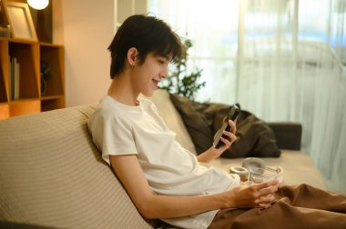 Gülümseyen Asyalı adam rahat koltukta akıllı telefon kullanıyor. Evde rahat bir anın tadını çıkarıyor..