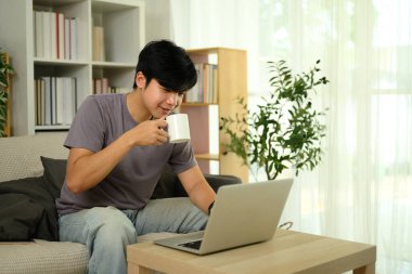 Gülümseyen Asyalı adam kahve içiyor ve laptopuyla oturma odasında çalışıyor..