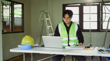 Mühendis proje planlarını gözden geçiriyor ve çalışma masasında dizüstü bilgisayar kullanıyor..