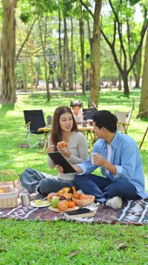 Mutlu aile ormanda kamp yapıyor, yiyecekleri paylaşıyor ve battaniyenin üzerinde dijital tablet kullanıyor..