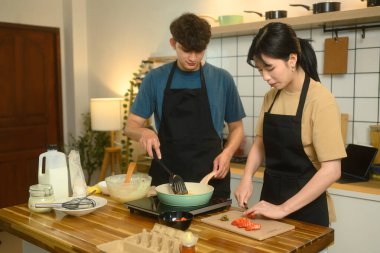Önlüklü bir çift kahvaltı hazırlıyor, krep yapıyor ve mutfakta taze meyve kesiyor..