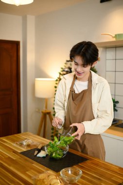 Önlüklü gülümseyen erkek modern bir mutfakta taze yeşil salata hazırlıyor..