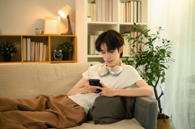 Genç Asyalı bir adam kanepede uzanıyor ve oturma odasında akıllı telefon kullanıyor..