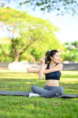 Genç bir kadın yoga minderinde oturuyor ve parkta egzersiz yaparken kolunu uzatıyor..