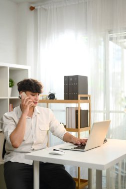 Kafkas genç girişimci iş yerinde cep telefonuyla konuşurken dizüstü bilgisayarını kullanıyor.