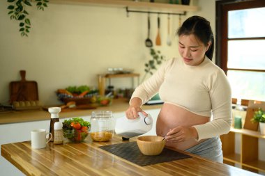 Hamile kadın sağlıklı bir kahvaltı hazırlıyor. Parlak bir mutfakta mısır gevreği ve sütle..