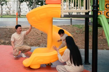 Mutlu ebeveynler güneşli bir günde küçük çocuklarıyla oyun parkında oynuyorlar...