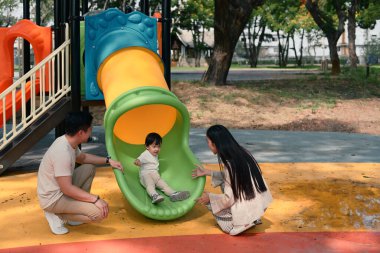 Mutlu ebeveynler güneşli bir günde küçük çocuklarıyla oyun parkında oynuyorlar...