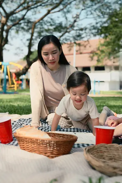 Anne ve küçük çocuk parkta piknik battaniyesinde dinleniyorlar, güneşli bir günün tadını çıkarıyorlar..