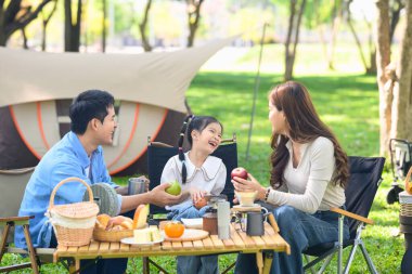 Ebeveynler ve neşeli kız doğadaki aile kamp gezisi sırasında rahatça dışarıda yemek yiyorlar..