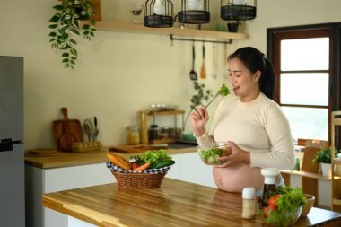 Gülümseyen hamile kadın mutfakta taze sebze salatası yiyor. Sağlıklı yaşam tarzı kavramı.