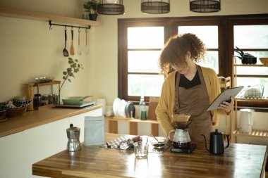 Genç bir kadın kahveyle demleniyor ve şık bir mutfakta dijital tablet kullanıyor..