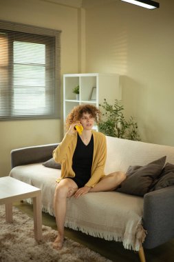 Kıvırcık saçlı kadın oturma odasında cep telefonuyla konuşuyor..