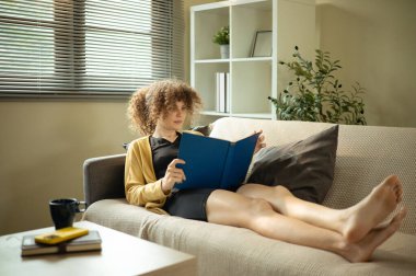 Kıvırcık saçlı sıradan bir kadın kanepede kitap okuyor..