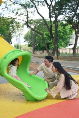 Anne ve baba, çocukları renkli bir oyun parkı kaydırağından inerken heyecanla gülümsüyor..