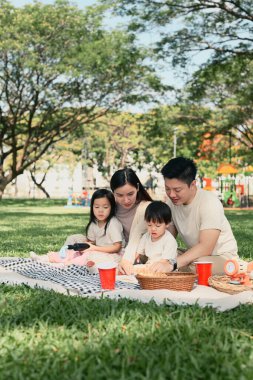 Ebeveynler ve iki çocuk yeşil bir parkta hafta sonu pikniği sırasında keyifli bir açık hava yemeğini paylaşıyorlar..