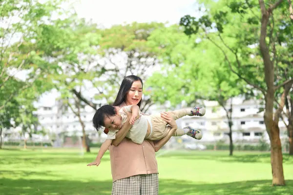 Anne, parlak ve güneşli yeşil bir parkta uçuyormuş gibi yaparak çocuğunu sevinçle havaya kaldırıyor..