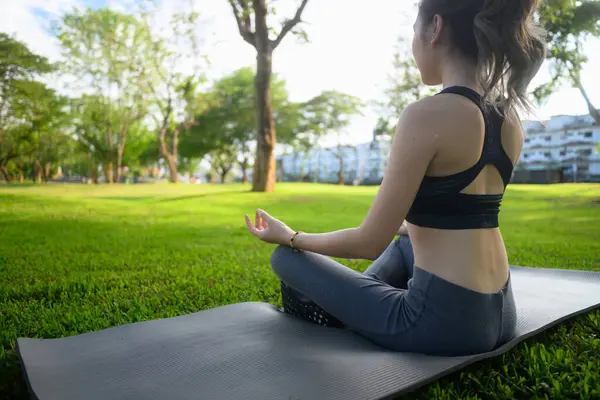 Huzurlu yeşil bir parktaki yoga minderinde Lotus pozisyonunda meditasyon yapan bir kadının arka görüntüsü..
