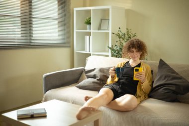 Kafkasyalı rahat bir kadın evdeki kanepede oturmuş elinde kupayla telefonunu kontrol ediyor..