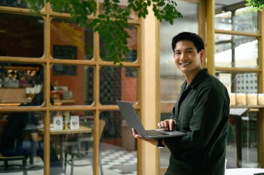 Elinde dizüstü bilgisayarla dışarıda duran gülümseyen genç bir adam..