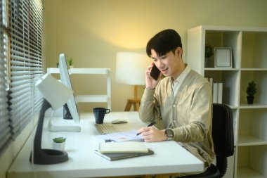 Modern bir masada notlar yazarken evde çalışan Asyalı genç bir adam telefonla konuşuyor..