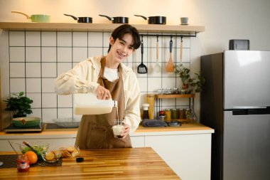 Neşeli genç adam modern bir mutfakta duran bardağa taze süt döker..