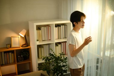 Genç adam pencerenin kenarında duruyor, rahat bir evde sabah güneşinin tadını çıkarıyor..