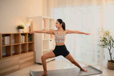 Genç bir kadın, parlak bir oturma odasında 2. Savaşçı yoga pozu veriyor..