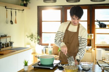 Önlüklü gülümseyen adam sıcak, modern mutfakta ocağın üstünde erişte hazırlıyor..