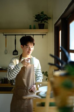 Önlüklü bir adam pencerenin kenarında duruyor, güneşli bir mutfakta sıcak bir içkinin tadını çıkarıyor..