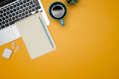 Dizüstü bilgisayarı, defteri, kahve fincanı ve sarı arka planda kablosuz kulaklıkları olan yaratıcı çalışma alanı.