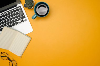 Sarı arka planda dizüstü bilgisayarı, defteri, gözlüğü ve kahve fincanı olan yaratıcı çalışma alanı.