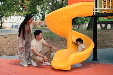 Mutlu ebeveynler, çocuklarının renkli bir kaydırağın kayışını izleyip alkışlıyorlar..