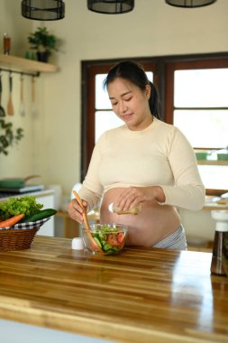 Gülümseyen hamile kadın cam bir kase sebze salatasına sos ekliyor..