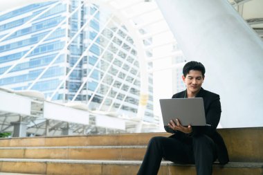 Resmi giyinmiş iş adamı, gökdelenlerle çevrili merdivenlerde otururken dizüstü bilgisayar kullanıyor..