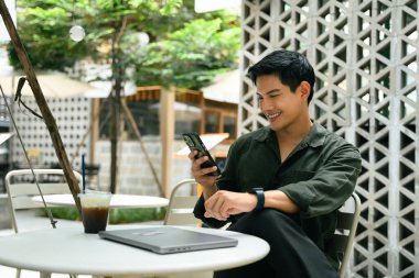 Gülümseyen adam açık bir kafede rahatlıyor ve kapalı bir dizüstü bilgisayarın yanında akıllı telefon kullanıyor..