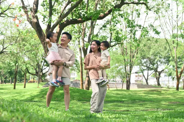 Anne ve baba çocuklarını kucağında tutuyorlar. Manzaralı bir parkta yürüyorlar..