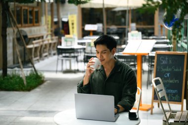 Laptop kullanan ve rahatlamış bir çalışma alanında kahve yudumlayan yakışıklı bir serbest çalışan..