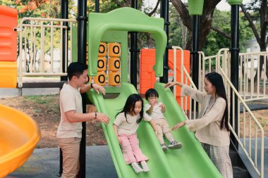 Ebeveynler, güneşli bir parkta iki küçük çocuğun oyun parkında kaymaktan zevk almasına sevinir..