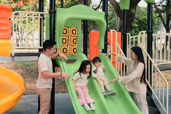 Ebeveynler, güneşli bir parkta iki küçük çocuğun oyun parkında kaymaktan zevk almasına sevinir..